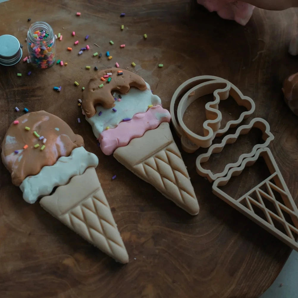 Ice cream cone and cookie shaped cookies on a wooden board with sprinkles and an ice cream scoop.
